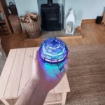 A hand holding a glowing, spherical Leteča Dron Žogica – Magični Player za Vse Generacije! v prijetni dnevni sobi. V sobi je lesena klubska mizica, kamin s pečjo in regal, ki ga krasijo okvirji za slike in okraski z motivom žogice.