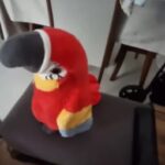 Plišasta igrača papiga v živo rdečih, rumenih in sivih barvah sedi na oblazinjenem stolu. Ozadje prikazuje del mize in stolov, s tkanino, prevlečeno čez en stol.