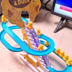Igralna steza z nizom modrih toboganov z majhnimi rumenimi figurami račk, ki plezajo in se spuščajo. Napis na vrhu se glasi "Small & Duck." V ozadju je viden računalniški monitor.
