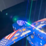 Roka drži GlideFly Penasto Letalo, ki prikazuje ameriško zastavo in zvezde. Igrača je osvetljena s pisanimi lučkami, ki ustvarjajo črte na temnem ozadju.